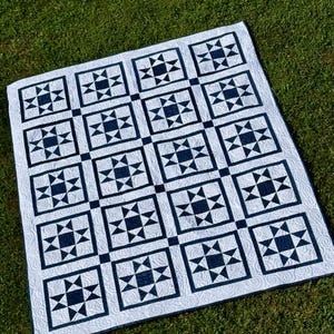 May include: A white and navy blue quilt with a repeating star pattern. The quilt is made of squares with a navy blue star design in the center of each square. The quilt is lying on green grass.