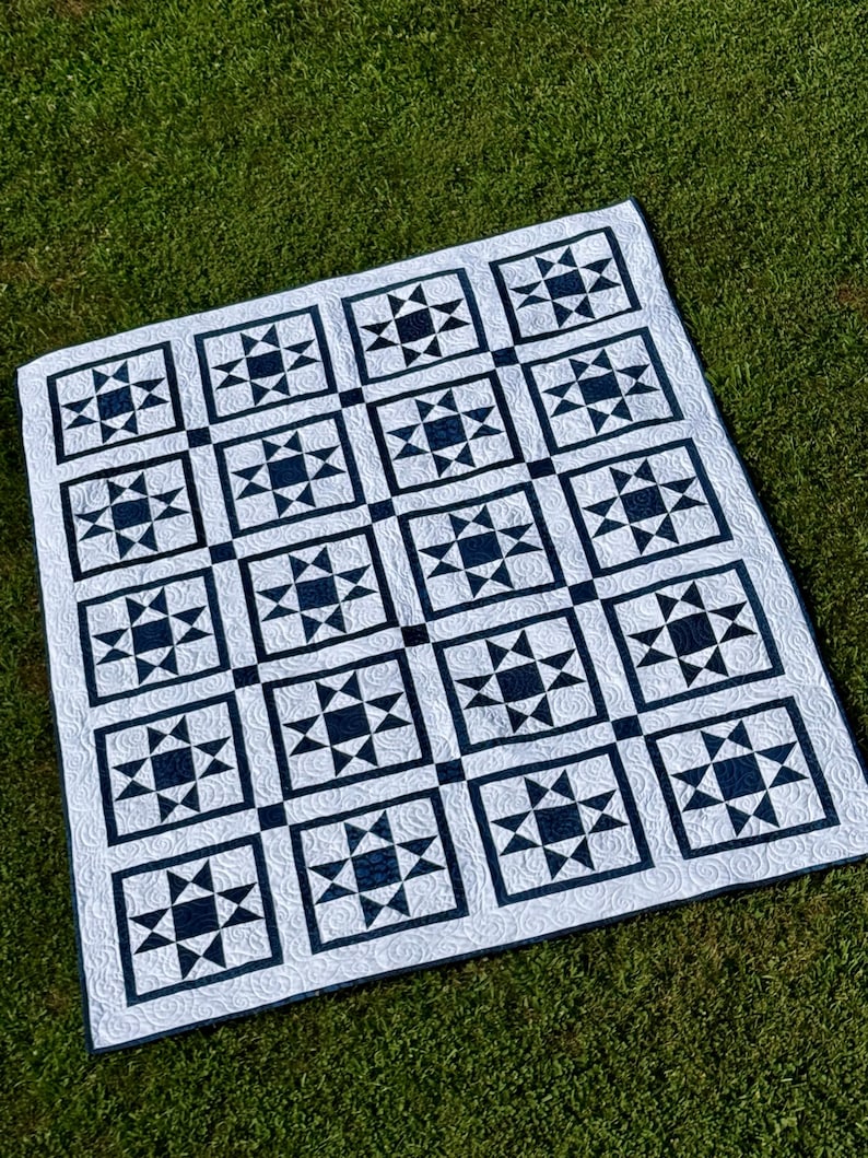 May include: A white and navy blue quilt with a repeating star pattern. The quilt is made of squares with a navy blue star design in the center of each square. The quilt is lying on green grass.