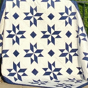 Op de afbeelding: Een wit quilt met een donkerblauw sterrenpatroon. Het quilt is gemaakt van herhalende stervormen met een ruit in het midden van elke ster.