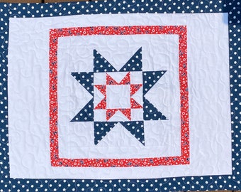 May include: A rectangular quilt featuring a central star pattern in navy blue with white polka dots and red fabric with small blue anchor prints. The design is framed by a solid white border and an outer navy blue border with white polka dots.