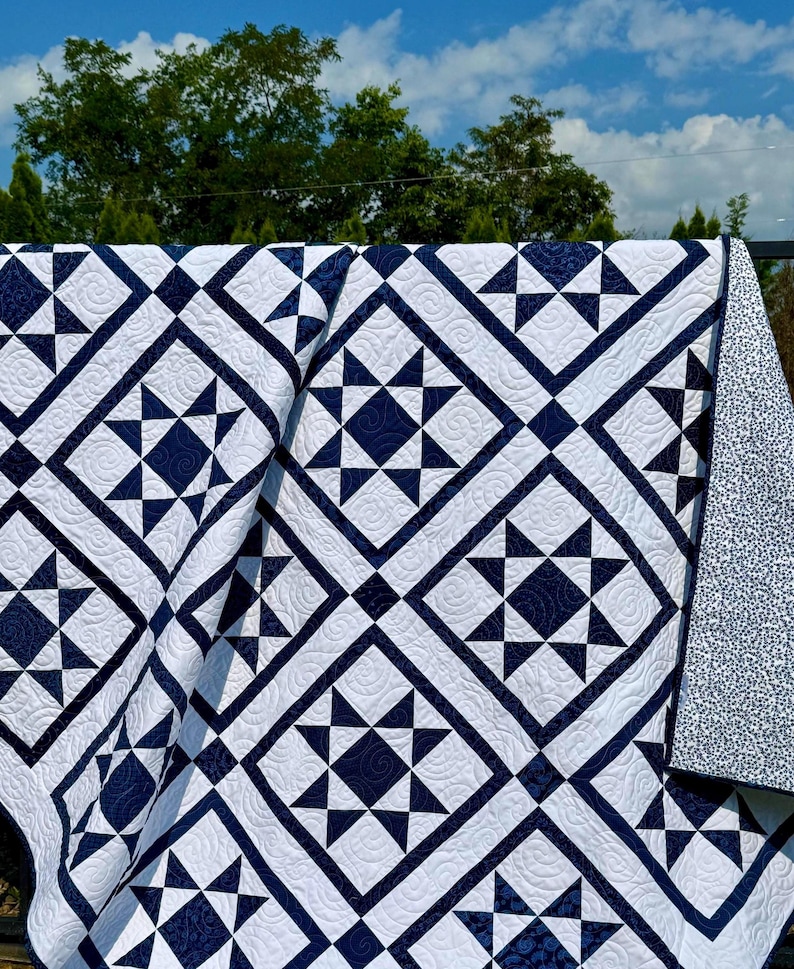 May include: A navy blue and white quilt with a star pattern. The quilt features a repeating geometric design of navy blue stars and diamonds on a white background. The quilt is draped over a railing, with a blue sky and trees in the background.