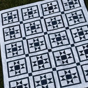 May include: A white and navy blue quilt with a star pattern. The quilt features a repeating design of navy blue stars within square blocks, separated by black lines. The background is white with a subtle, textured pattern.