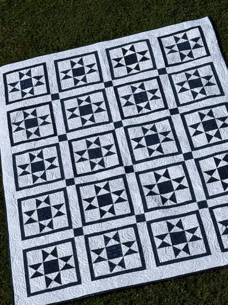 May include: A white and navy blue quilt with a star pattern. The quilt features a repeating design of navy blue stars within square blocks, separated by black lines. The background is white with a subtle, textured pattern.