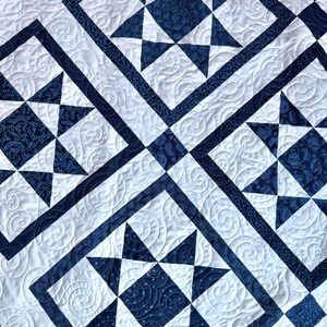 May include: A close-up of a quilted blanket featuring a repeating star pattern. The quilt has a white background with navy blue star shapes and borders. The stars are made of solid and patterned fabric, with intricate stitching.