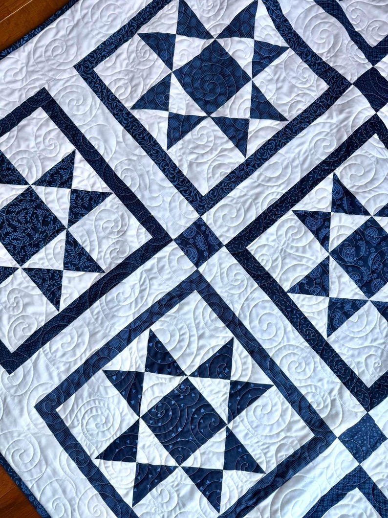 May include: A close-up of a quilted blanket featuring a repeating star pattern. The quilt has a white background with navy blue star shapes and borders. The stars are made of solid and patterned fabric, with intricate stitching.