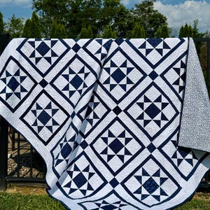 May include: A white and navy blue quilt with a geometric star pattern. The quilt is draped over a black metal fence, with a grassy lawn and trees in the background. The quilt's design features alternating squares and star shapes.