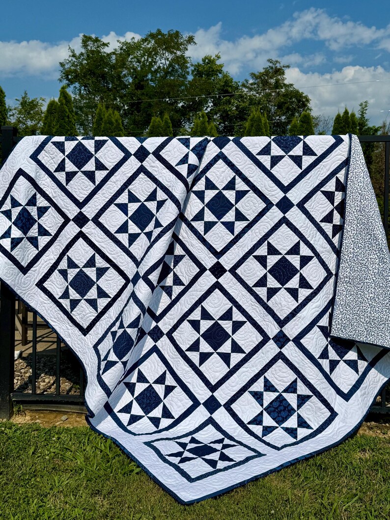 May include: A white and navy blue quilt with a geometric star pattern. The quilt is draped over a black metal fence, with a grassy lawn and trees in the background. The quilt's design features alternating squares and star shapes.