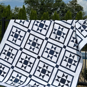 May include: A white and navy blue quilt with a star pattern. The quilt features a repeating design of navy blue stars within square frames, set against a white background. The quilt is draped over a black fence in an outdoor setting.