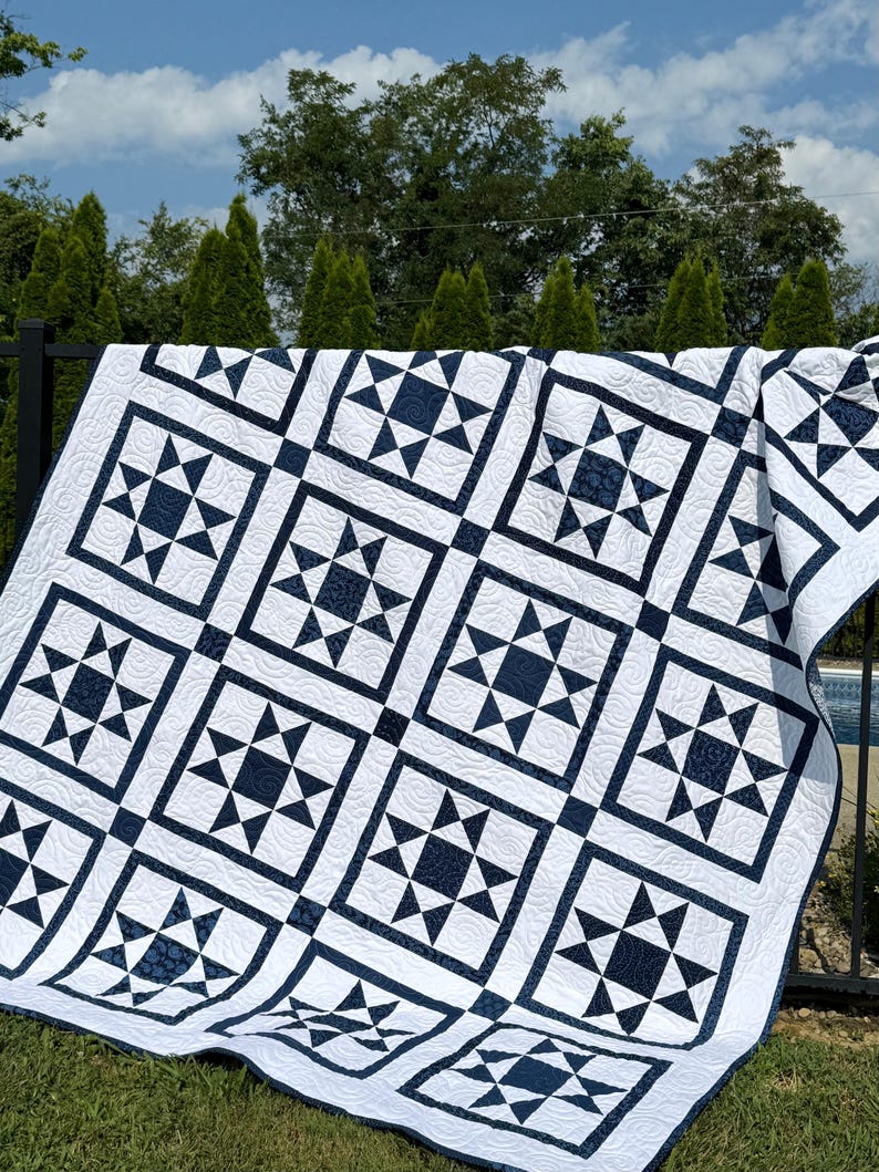 May include: A white and navy blue quilt with a star pattern. The quilt features a repeating design of navy blue stars within square frames, set against a white background. The quilt is draped over a black fence in an outdoor setting.