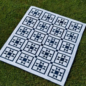 May include: A white quilt with a navy blue star pattern laid out on green grass. The quilt features a repeating design of square blocks, each containing a star and triangle motif. The quilt is a handmade item.