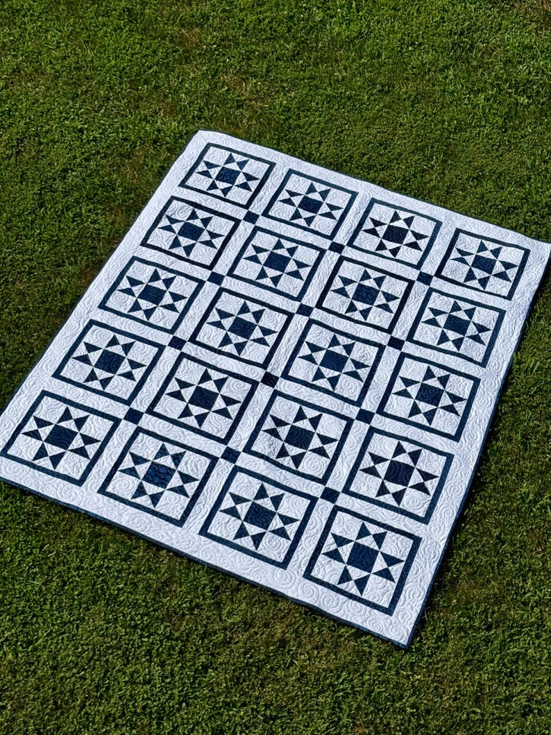May include: A white quilt with a navy blue star pattern laid out on green grass. The quilt features a repeating design of square blocks, each containing a star and triangle motif. The quilt is a handmade item.