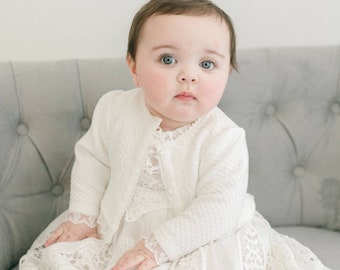 Puede incluir: Un vestido color crema de manga larga con intrincados detalles de encaje en la falda y el corpiño, combinado con un cárdigan a juego de textura acolchada que tiene un cierre de un solo botón en el cuello y encaje en los puños de las mangas.