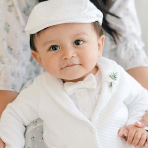 May include: A baby boy wearing a white suit with a bow tie and a white cap. He is holding a small bouquet of flowers.