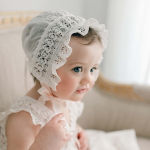 May include: A white lace bonnet with a ruffled edge. The bonnet is worn by a baby with blue eyes.