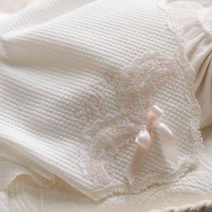Può includere: Una coperta per neonati bianca e strutturata con un motivo a nido d'ape. La coperta presenta un delicato bordo in pizzo in una morbida tonalità rosa, con il nome "Sophia" ricamato con filo rosa abbinato. Un fiocco di raso aggiunge un tocco finale.