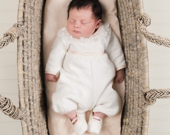 Puede incluir: Un bebé durmiendo con un mono acolchado blanco con ribete de encaje y zapatos blancos, está acostado en una cesta de mimbre.