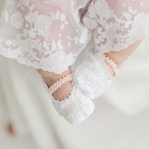 May include: A pair of white lace baby booties with pink satin ribbon accents. The booties are worn by a baby wearing a white lace dress.