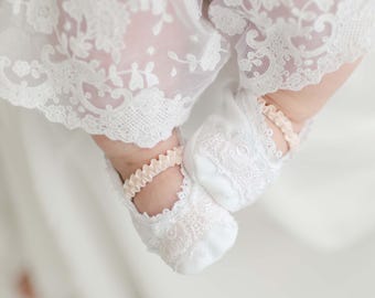 Könnte beinhalten: Ein Paar weiße Spitzen-Babyschuhe mit rosa Satinband-Akzenten. Die Schuhe werden von einem Baby getragen, das ein weißes Spitzenkleid trägt.