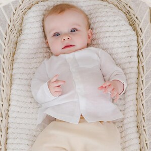 Puede incluir: Un bebé con una camisa blanca abotonada y pantalones beige yace en una cuna de mimbre blanca. El bebé tiene la piel clara, ojos azules y mira hacia arriba. La cuna está forrada con una manta blanca texturizada.