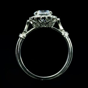 K&ouml;nnte beinhalten: Ein silberner Ring mit einem gro&szlig;en, runden, klaren Diamanten, der in einem Halo aus kleineren Diamanten gefasst ist. Der Ring hat ein zartes, filigranes Design.