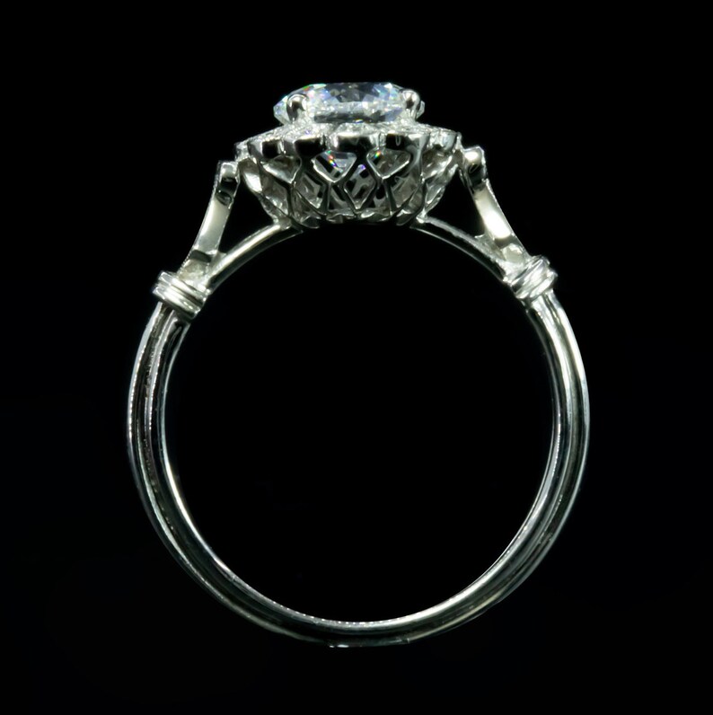 K&ouml;nnte beinhalten: Ein silberner Ring mit einem gro&szlig;en, runden, klaren Diamanten, der in einem Halo aus kleineren Diamanten gefasst ist. Der Ring hat ein zartes, filigranes Design.