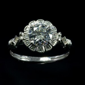 K&ouml;nnte beinhalten: Ein silberner Verlobungsring mit einem gro&szlig;en, runden, klaren Diamanten, der von kleineren Diamanten in einem floralen Design umgeben ist.