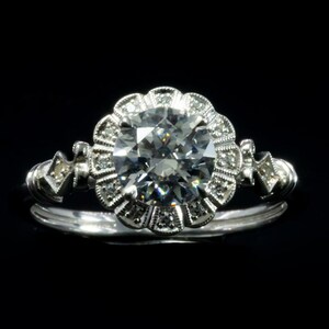 K&ouml;nnte beinhalten: Ein silberner Verlobungsring mit einem gro&szlig;en, runden, klaren Diamanten, der von einem Heiligenschein aus kleineren Diamanten umgeben ist. Die Band ist auf jeder Seite mit kleineren Diamanten verziert.