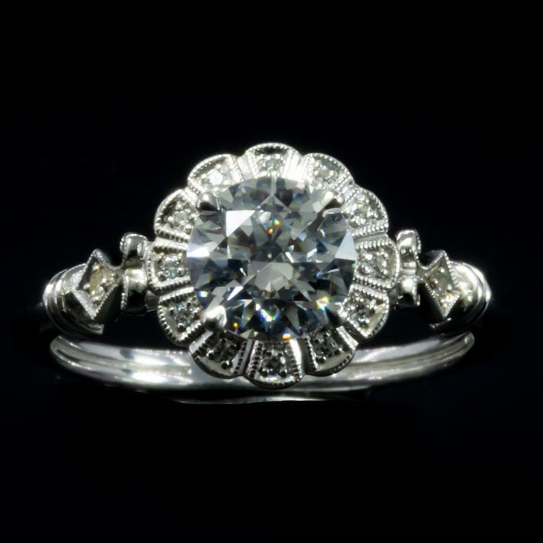 K&ouml;nnte beinhalten: Ein silberner Verlobungsring mit einem gro&szlig;en, runden, klaren Diamanten, der von einem Heiligenschein aus kleineren Diamanten umgeben ist. Die Band ist auf jeder Seite mit kleineren Diamanten verziert.