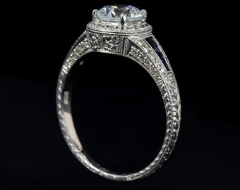 May include: A silver-toned ring featuring a large round center stone surrounded by a halo of small clear stones. The band is decorated with intricate engravings and features a row of small, deep blue rectangular stones along the side shank.