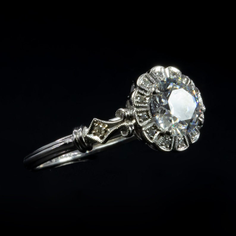 K&ouml;nnte beinhalten: Ein silberner Verlobungsring mit einem gro&szlig;en, runden, klaren Diamanten, der in einem floralen Heiligenschein gefasst ist. Der Ring hat eine zarte, verzierte Band.