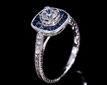 May include: A close-up of a silver ring with a large, clear center stone surrounded by smaller blue stones. The ring has intricate detailing on the band and around the stones, set against a black background.