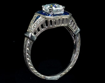 May include: A silver ring with a large, clear, square-shaped center stone surrounded by smaller blue stones. The band is intricately designed with a filigree pattern.