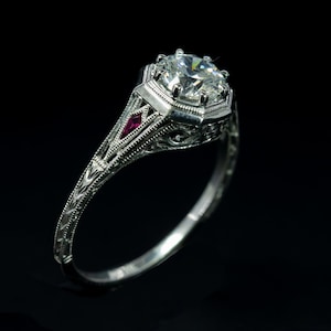 Op de afbeelding: Een zilveren ring met een grote, ronde, heldere diamant en een klein, rood robijn accent. De ring heeft een delicaat, filigraan ontwerp.