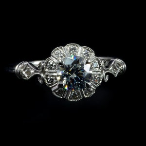 K&ouml;nnte beinhalten: Ein silberner Verlobungsring mit einem gro&szlig;en, runden, klaren Diamanten, der von kleineren Diamanten in einem floralen Design umgeben ist.