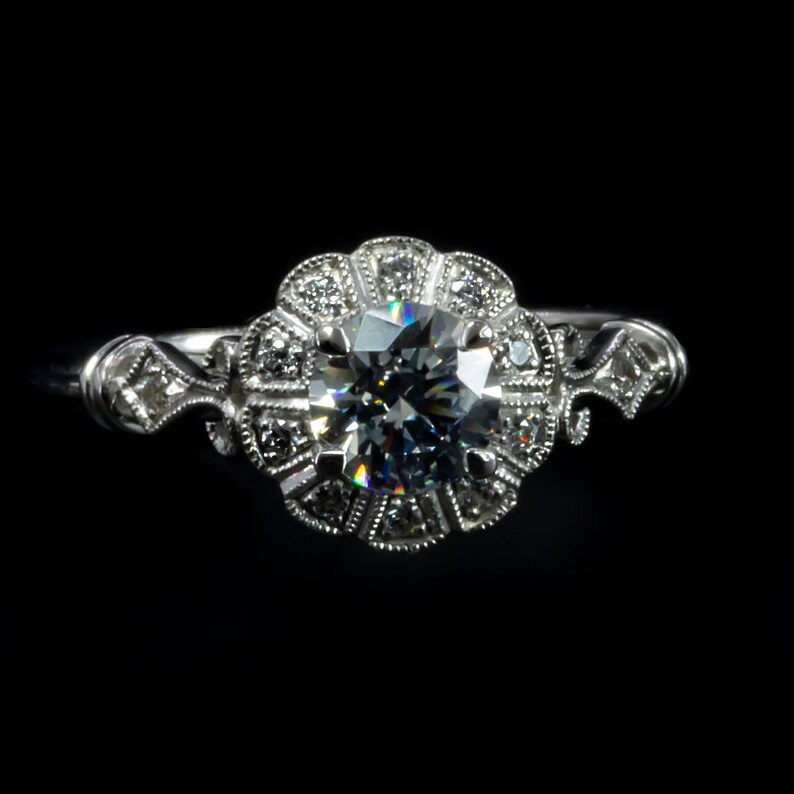 K&ouml;nnte beinhalten: Ein silberner Verlobungsring mit einem gro&szlig;en, runden, klaren Diamanten, der von kleineren Diamanten in einem floralen Design umgeben ist.