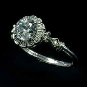 K&ouml;nnte beinhalten: Ein silberner Ring mit einem gro&szlig;en, runden, klaren Diamanten, der in einem floralen Design gefasst ist. Die Band ist d&uuml;nn und zart.