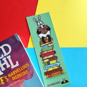 May include: A colourful bookmark featuring a cartoon sheep reading a book, standing on a stack of books with titles like "Drac-ewe-la" and "Baaary Potter". The bookmark has the text "eweniverse" at the bottom.