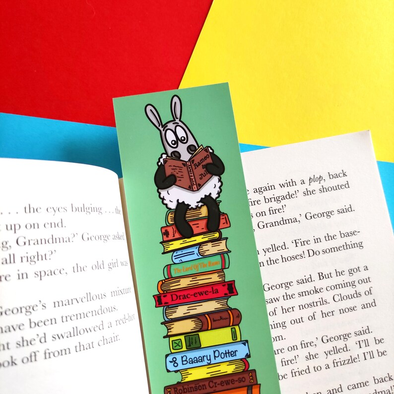 May include: A green bookmark featuring a cartoon sheep reading a book titled "Rameo and Juliet". The sheep is sitting on a stack of books with titles like "The Land of The Ban", "Drac-ewe-la", "8 Baaary Potter", and "Robinson Cr-ewe-so".