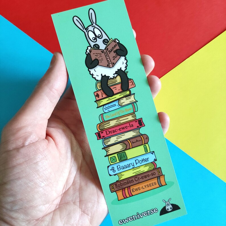 May include: A green bookmark featuring a cartoon sheep reading a book titled "Rameo Jul". The sheep is perched atop a stack of books with titles such as "The Lord of the Ram", "Drac-ewe-la", "8 Baaary Potter", "Robinson Cr-ewe-so", and "EWE-LYSEES". The bookmark is signed "eweniverse" at the bottom.