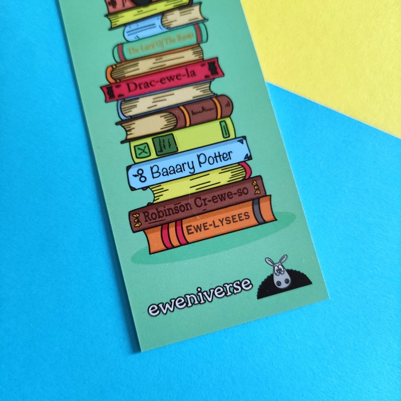 May include: A green bookmark featuring a stack of illustrated books with humorous titles such as "Drac-ewe-la" and "8 Baaary Potter". A small black and white donkey is peeking out from behind the books. The text "eweniverse" is printed at the bottom of the bookmark.