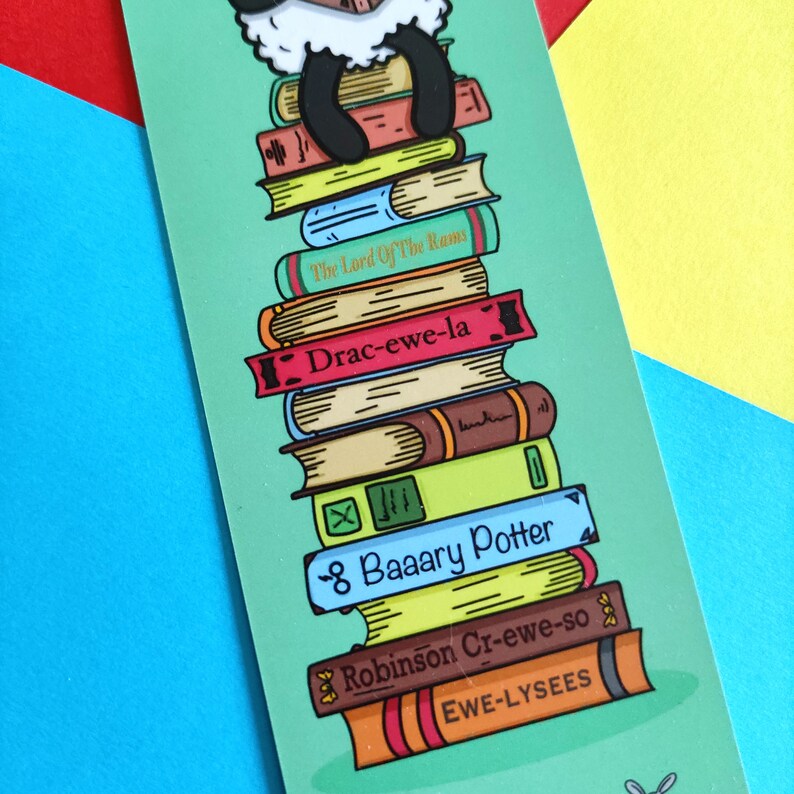 May include: A green bookmark featuring a white sheep perched atop a stack of books. The books have titles such as "The Lord Of The Rams", "Drac-ewe-la", "8 Baaary Potter", "Robinson Cr-ewe-so", and "EWE-LYSEES".