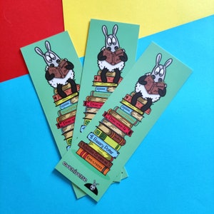 May include: Three green bookmarks featuring a cartoon sheep reading a book on a stack of books. The books have titles such as "The Lord of the Rings", "Drac-ewe-la", "8 Baaary Potter", and "Robinson Cr-ewe-so". The bookmarks are labelled "eweniverse" at the bottom.