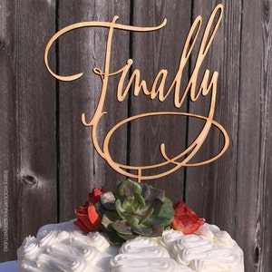 May include: A white cake with white frosting and a wooden cake topper that says "Finally" in cursive. The cake is decorated with red flowers and green succulents.