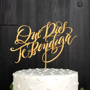 May include: A white cake with a textured surface and small white dots, decorated with green leaves. A wooden cake topper in elegant script reads "Que Dios Te Bendiga."