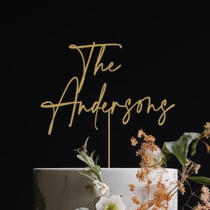 May include: A gold-colored cake topper featuring the script text The Andersons, supported by a thin vertical stake. The topper is positioned atop a white cake decorated with white flowers and dried foliage against a solid black background.