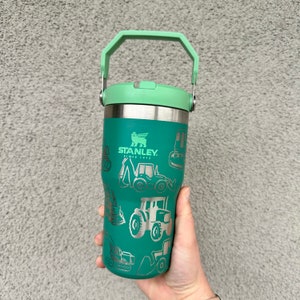May include: A green Stanley travel mug with a handle. The mug has a stainless steel exterior and is decorated with a pattern of construction vehicles. The text "STANLEY SINCE 1913" is printed on the mug.