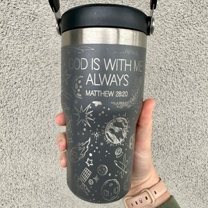 May include: A gray stainless steel travel mug with a black lid and a black handle. The mug has a white design of stars, planets, and other celestial objects. The text "GOD IS WITH ME ALWAYS" and "MATTHEW 28:20" is engraved on the mug.
