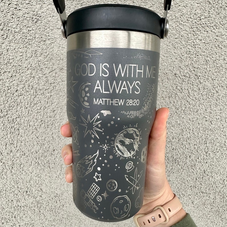 May include: A gray stainless steel travel mug with a black lid and a black handle. The mug has a white design of stars, planets, and other celestial objects. The text "GOD IS WITH ME ALWAYS" and "MATTHEW 28:20" is engraved on the mug.