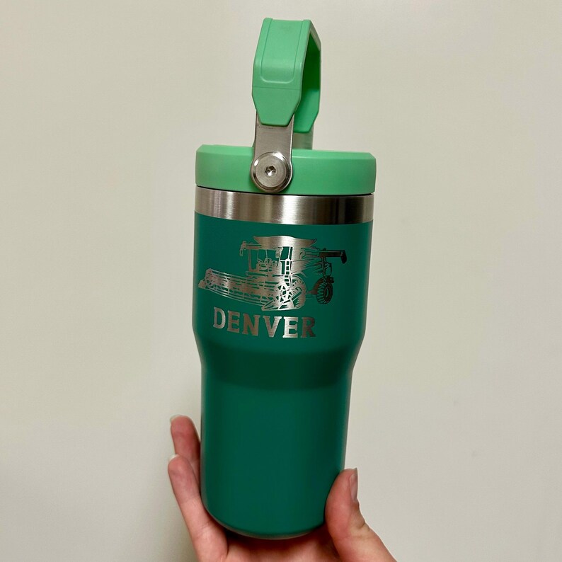May include: A green stainless steel travel mug with a green lid and a handle. The mug has a silver design of a combine harvester and the word "DENVER" on the side.