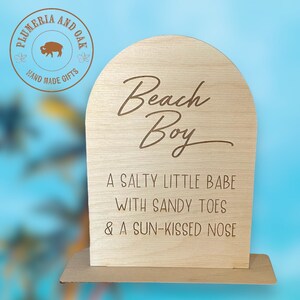 May include: Wooden sign with a light brown stain. The sign is shaped like an arch and has the text "Beach Boy" in a cursive font. Below the text is the phrase "A salty little babe with sandy toes & a sun-kissed nose".